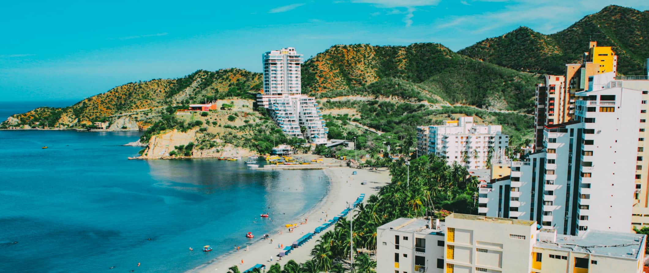 Vista do ar para a faixa de praia em Santa Marte, na Colômbia, com hotéis ao longo da costa e colinas ao fundo.