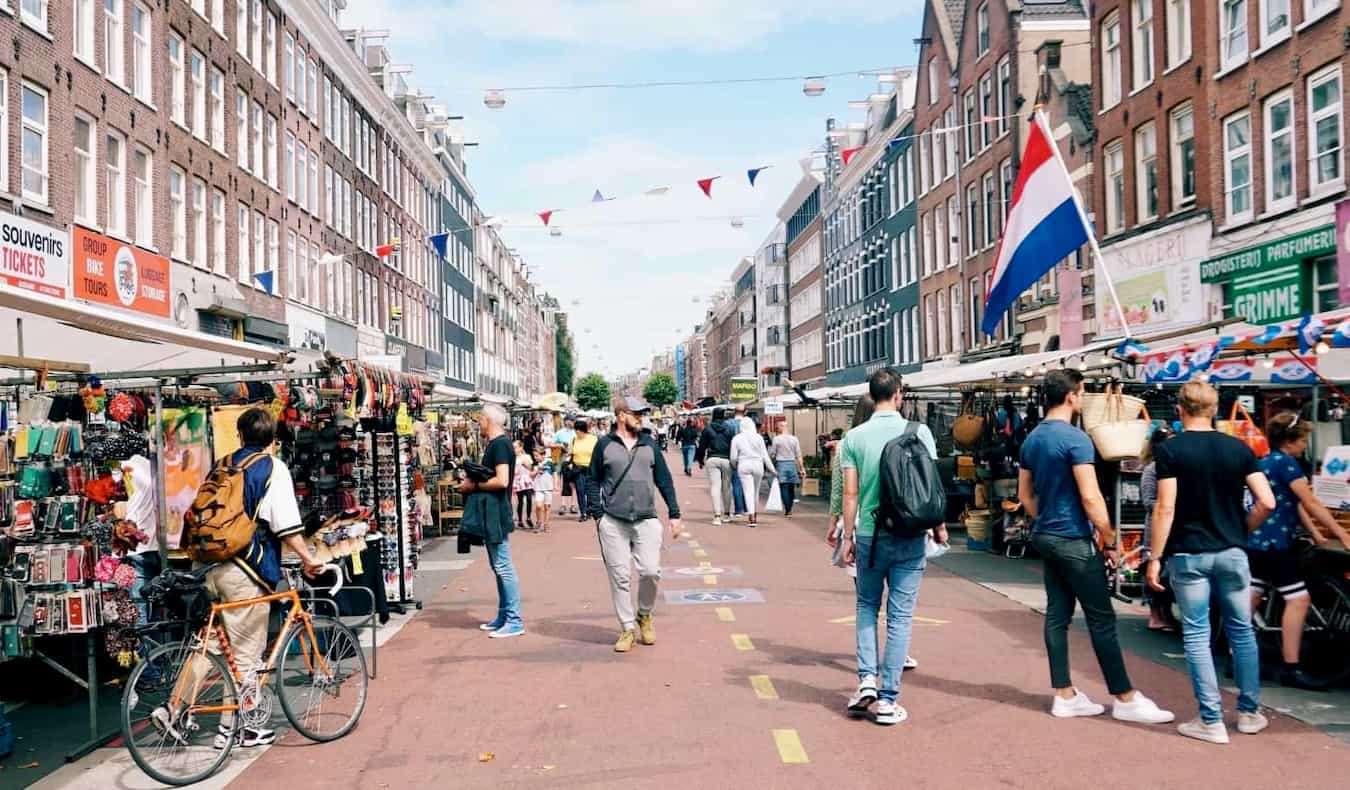 As pessoas andam ao longo de uma estreita rua movimentada em Sunny Amsterdã, olhando para pequenas lojas.