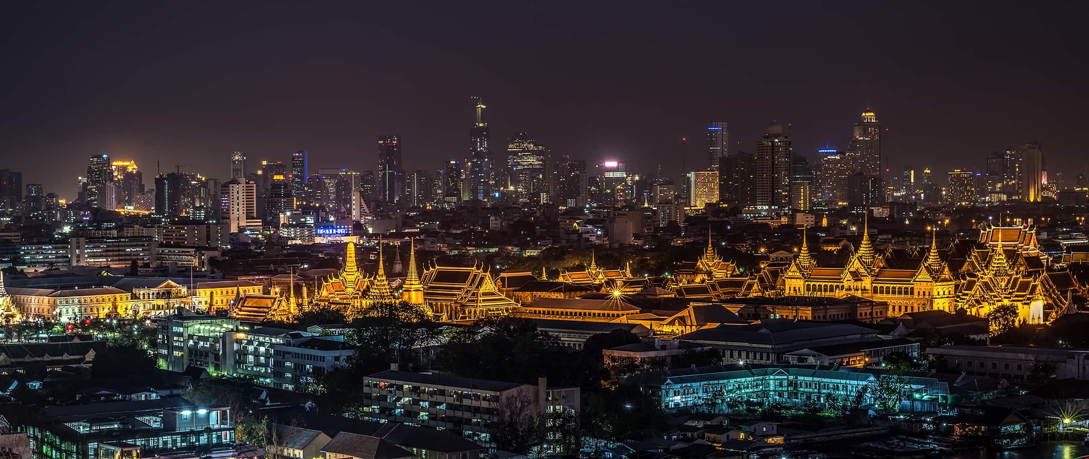 A linha do horizonte de Night Bangkok (Tailândia) com edifícios baixos em primeiro plano, o complexo do templo no centro e arranh a-céus modernos no fundo