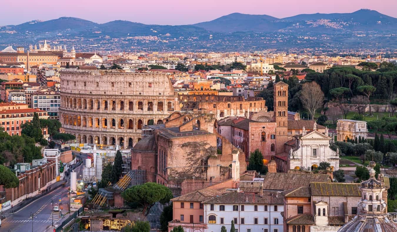 Horizonte de Roma, Itália, ao pôr do sol, com edifícios históricos, incluindo o Coliseu, em primeiro plano e montanhas ao fundo.