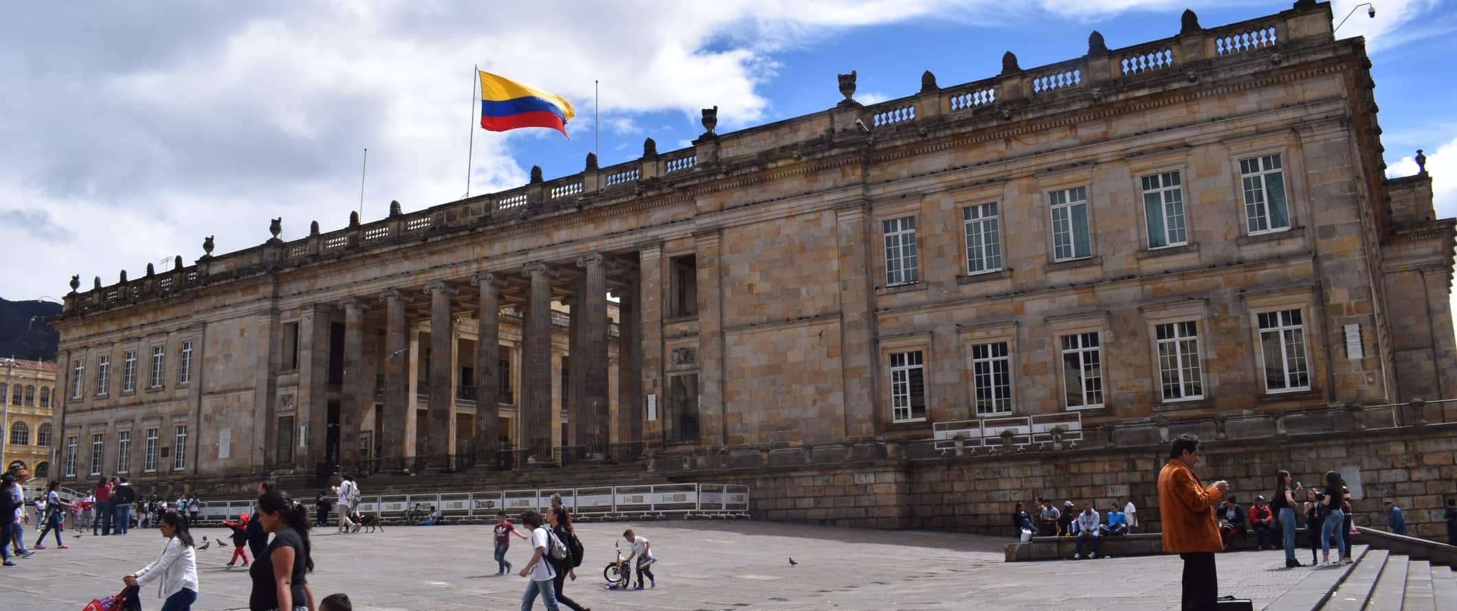 As pessoas andam em frente a um grande edifício neoclássico com muitas colunas e a bandeira colombiana, vibrando no telhado, em Bogotá, Colômbia