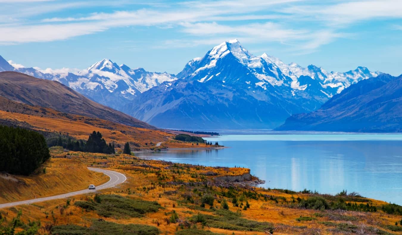 O microônibus cavalga ao longo de uma estrada sinuosa com um lago e montanhas ao fundo na Nova Zelândia.