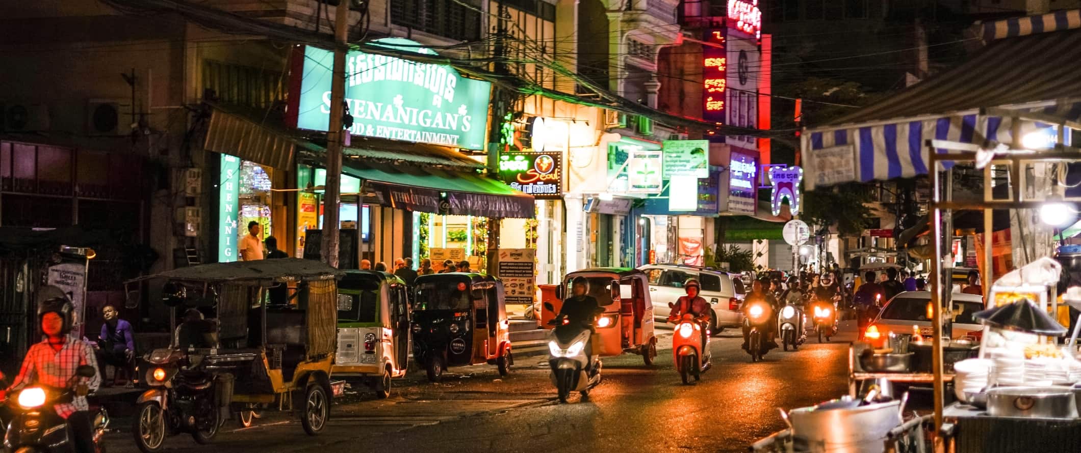 Ciclomotores, motocicletas e tuk-tuks circulam por uma rua iluminada à noite em Phnom Penh, capital do Camboja.