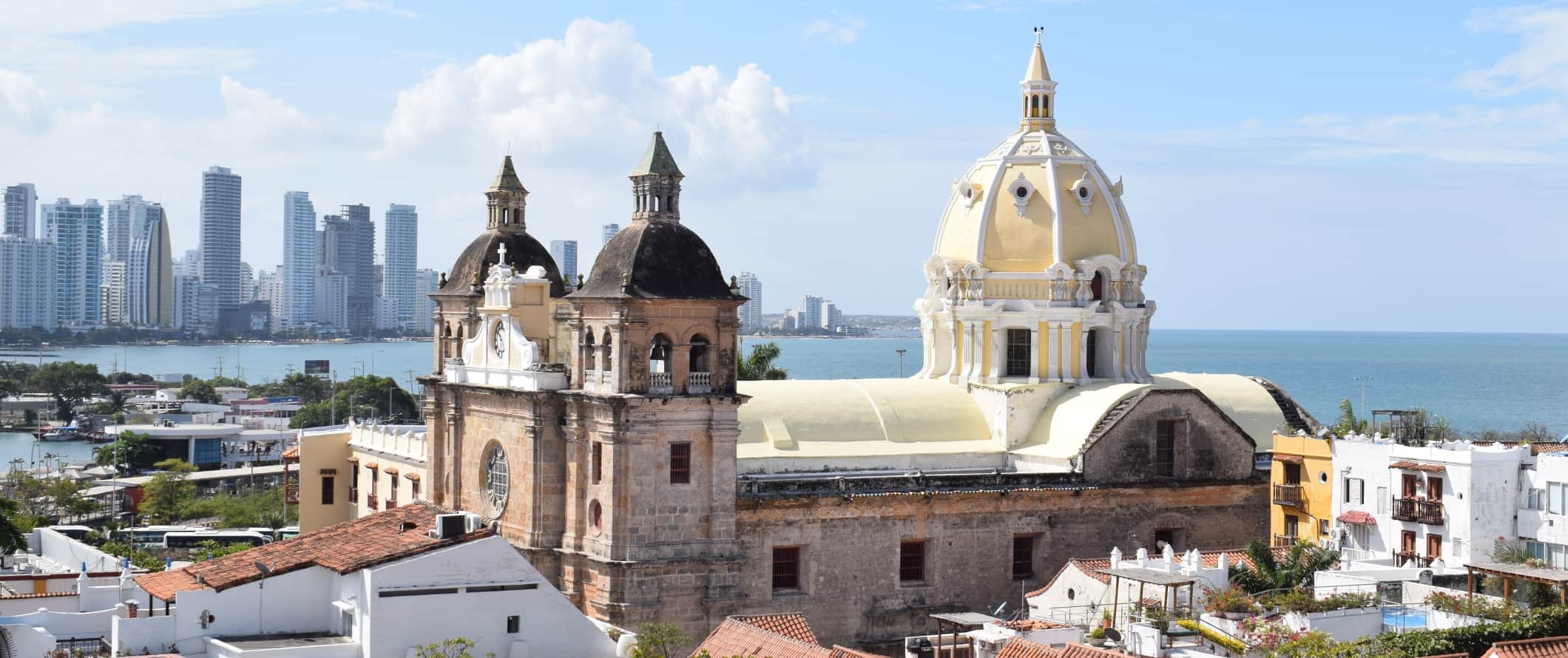 Vista da cidade de Cartagena, Colômbia, com uma grande igreja histórica de cúpula em primeiro plano e arranh a-céus modernos ao fundo.
