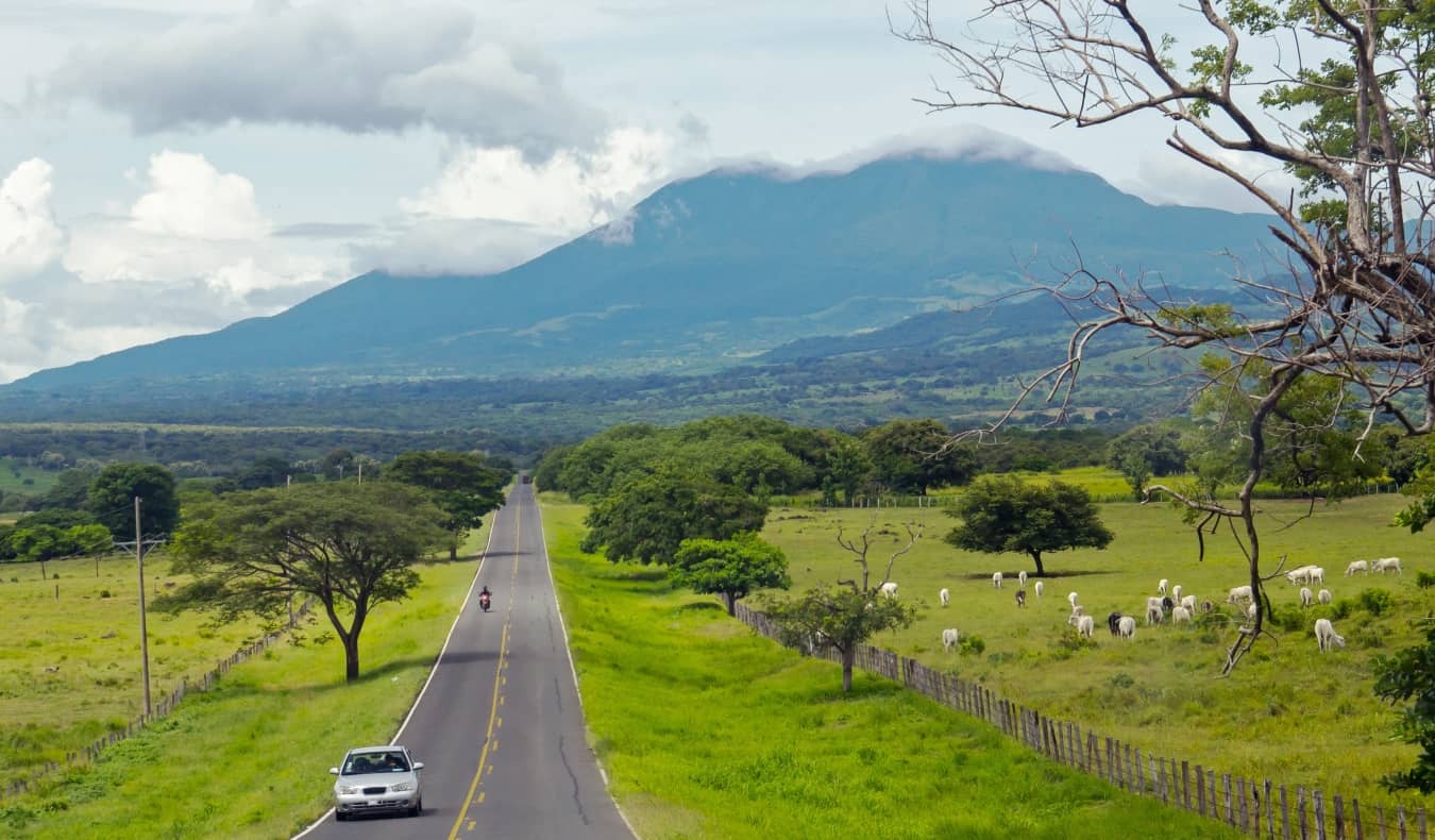 Carros viajam pela estrada para a Costa Rica com um vulcão ao fundo e vacas pastando ao longo das estradas