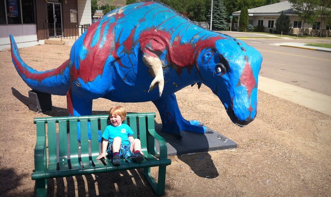 A criança chora, sentada em um banco perto da estátua azul de um dinossauro durante as férias