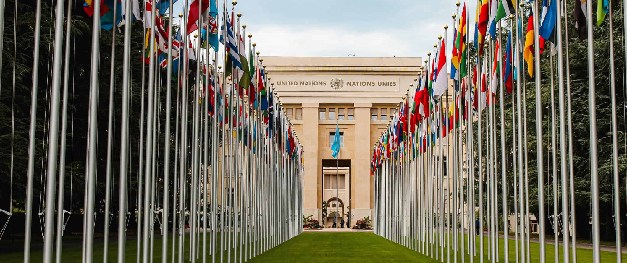 O prédio das Nações Unidas com duas fileiras de bandeiras de diferentes países do mundo à sua frente em Genebra, Suíça
