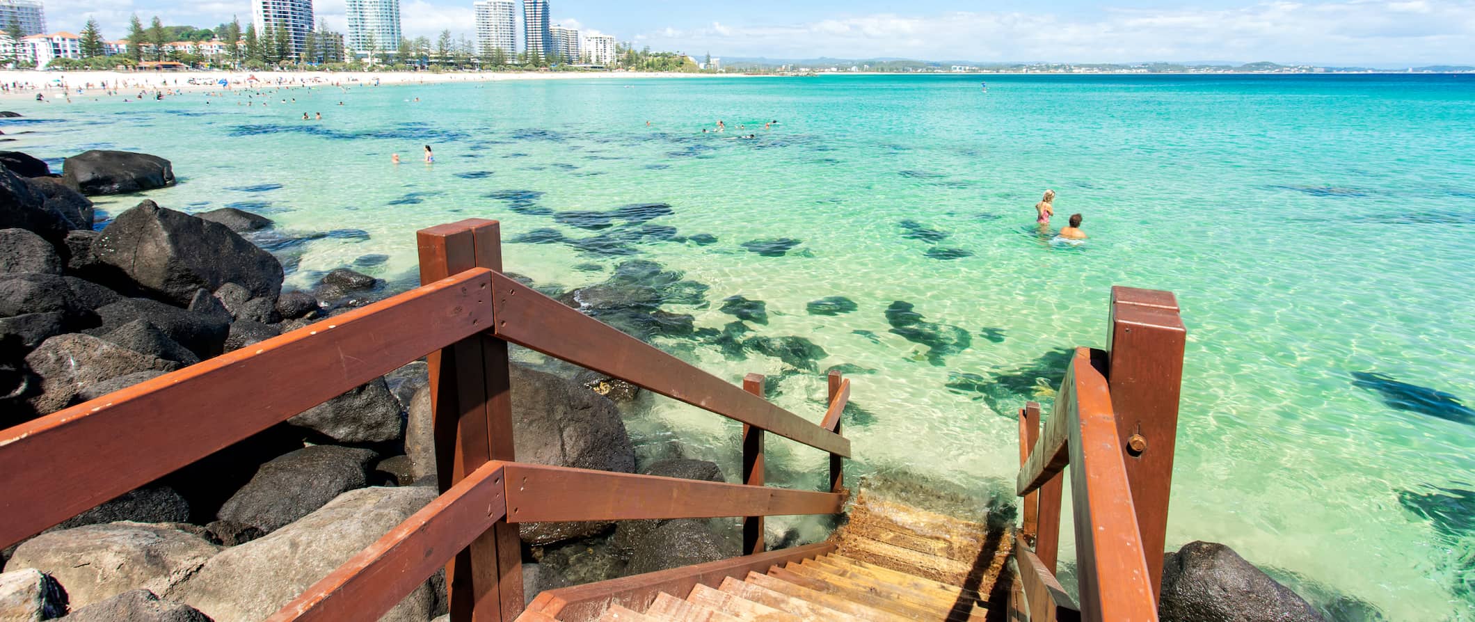 Pessoas nadam nas águas cristalinas de Gold Coast, na Austrália, perto de uma pequena escadaria de madeira com a cidade ao fundo.