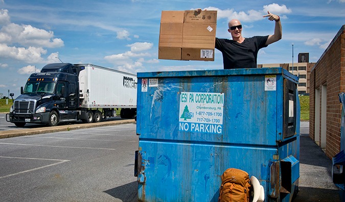 Um cara em um tanque de lixo segura um pedaço de papelão para seu sinal de carona, um caminhão passa