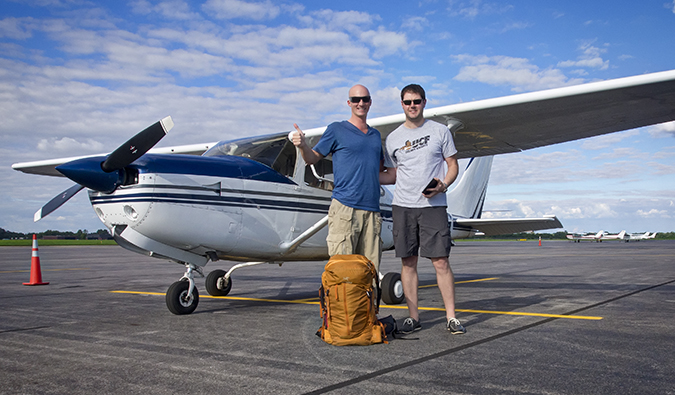 Dois caras posam na frente de um avião com uma mochila e polegar
