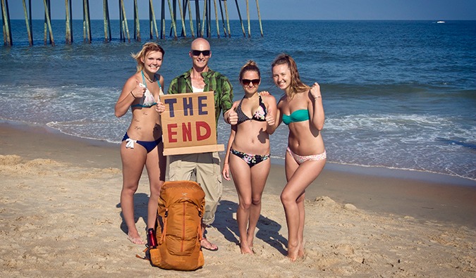Um homem fica na praia com 3 meninas em um biquíni e um sinal de papelão de um carona com a inscrição no fim