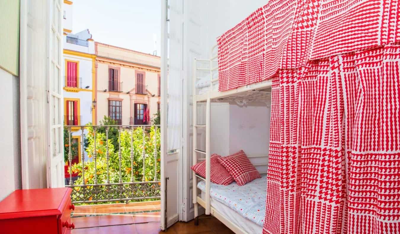Camas duplas com cortinas quadrilhas vermelhas, uma porta aberta para o terraço, com a qual os edifícios brilhantes de Sevilha, Espanha são visíveis ao fundo