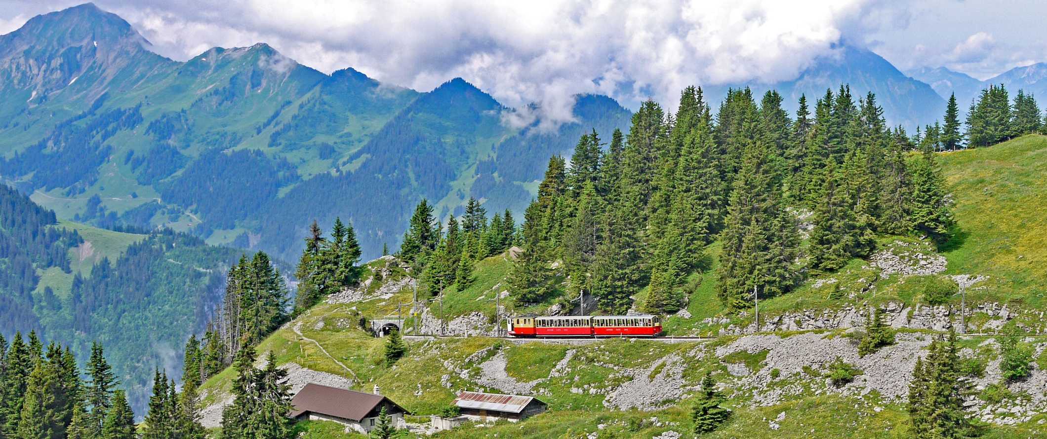O trem vermelho sobe ao longo de uma encosta íngreme com picos de montanha afiados ao fundo da ferrovia Jungfrayokh em Interlacten, na Suíça.