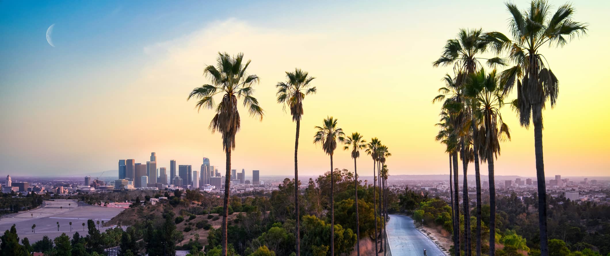 Los Angeles ao pôr do sol, com edifícios imponentes e palmeiras em primeiro plano.