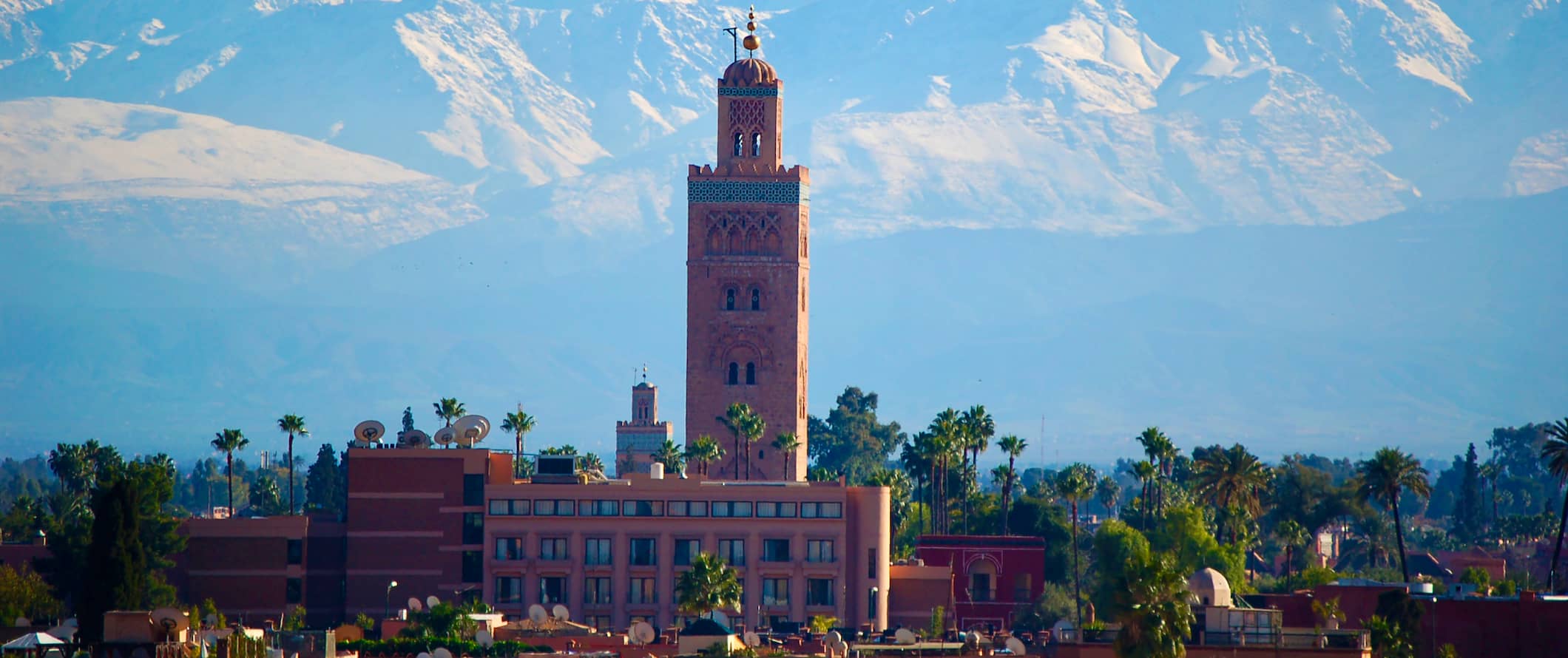 Vista da cidade de Marrakech, Marrocos, com torre em primeiro plano e montanhas deslumbrantes à distância