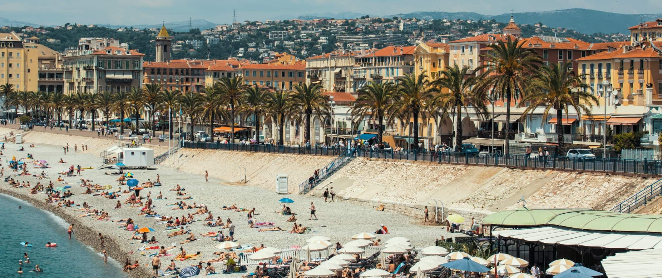 As pessoas deitam na praia em frente ao aterro sentado com palmeiras e, em segundo plano, a cidade de Nice, a França se eleva.