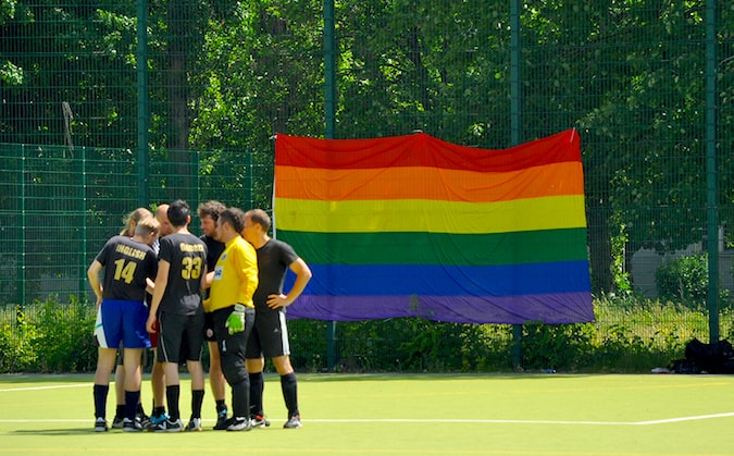 Um time de futebol fica em um campo com uma enorme bandeira de orgulho em segundo plano