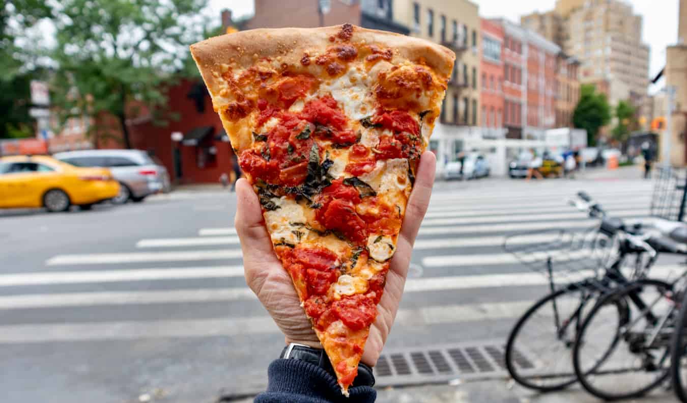 Mão segurando um pedaço de pizza em uma das ruas de Nova York