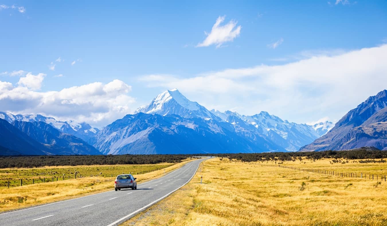 O carro viaja pela estrada com campos de ouro de ambos os lados, levando às montanhas nevadas na ilha sul da Nova Zelândia