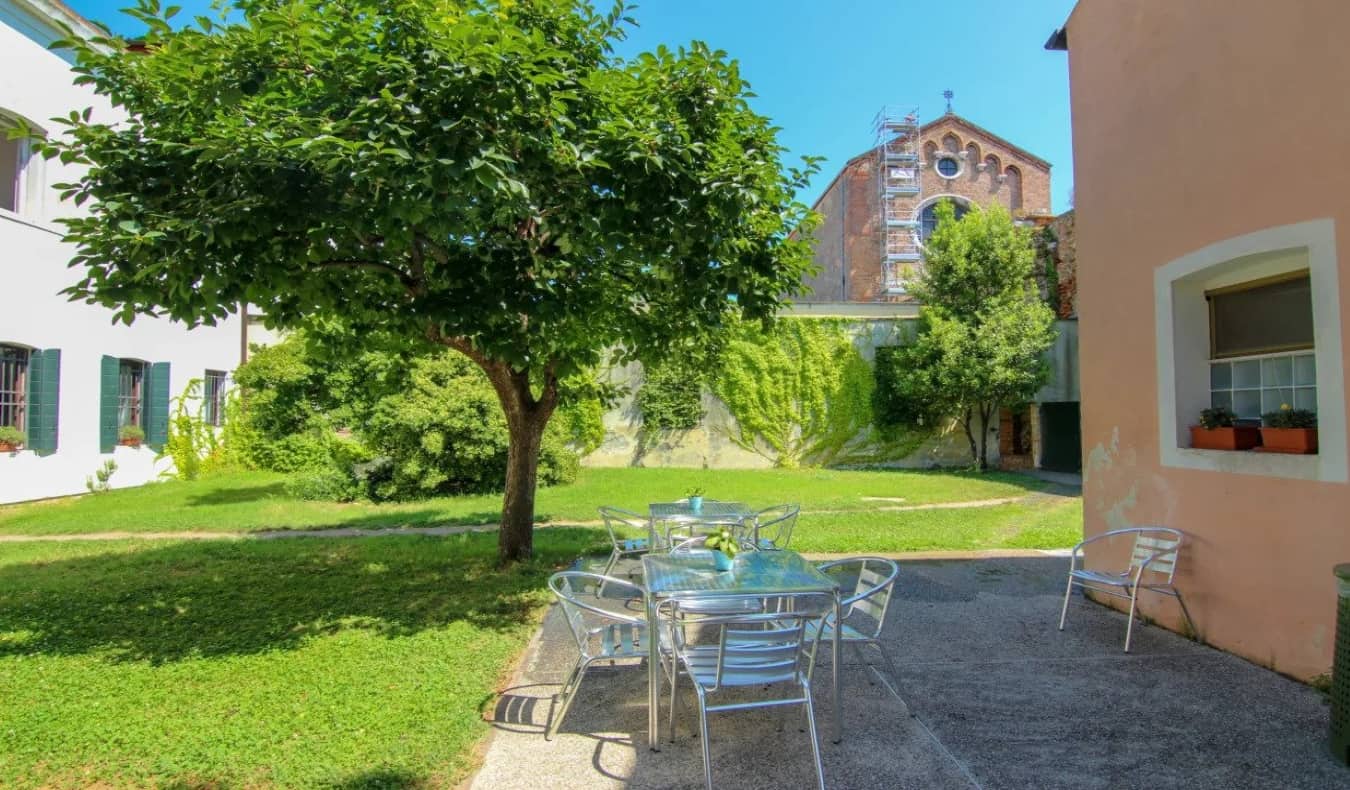 Um quintal traseiro fechado com árvores, mesas de metal no pátio e a igreja à distância no albergue Ostell S. Fosca em Veneza, Itália.