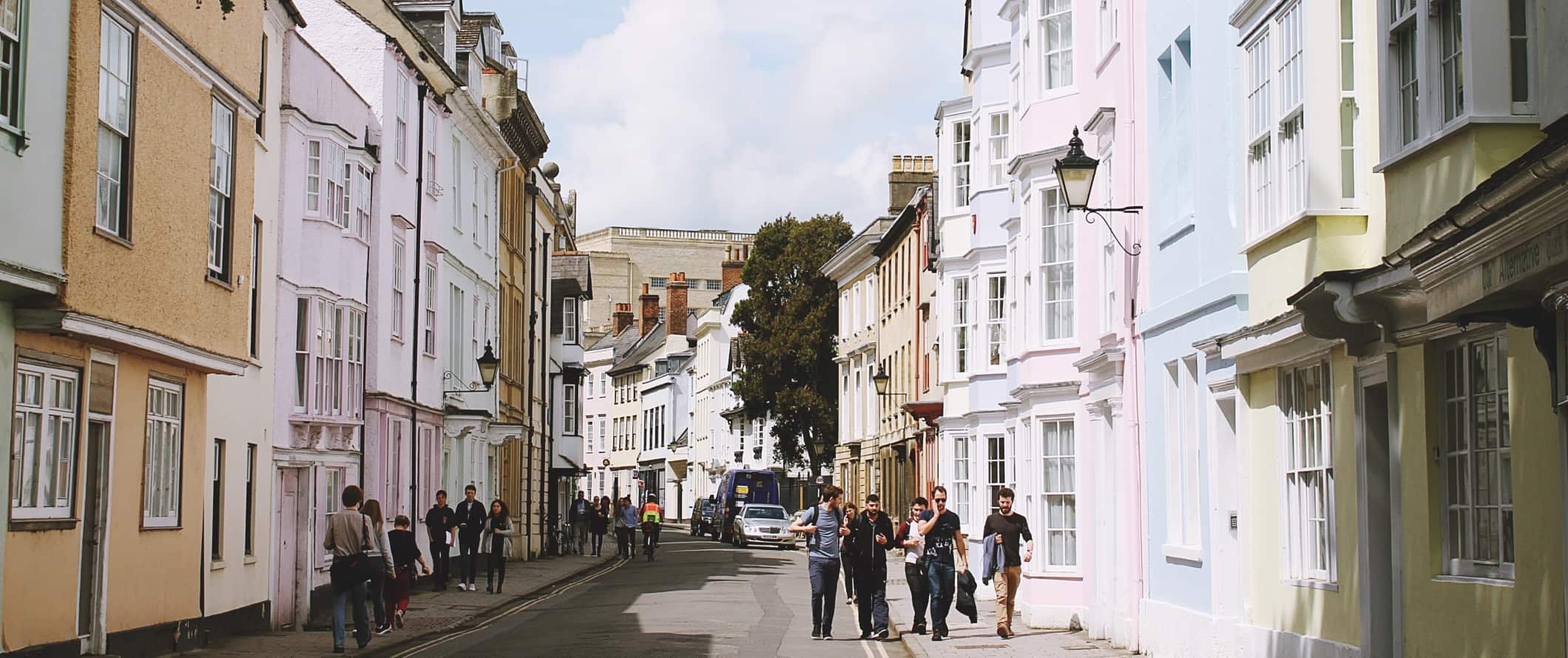 Pessoas caminham por uma rua repleta de casas em tons pastéis em Oxford, Inglaterra.