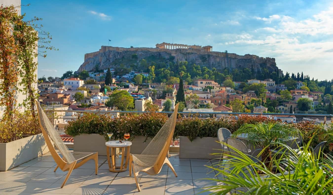Espreguiçadeiras de madeira em frente a uma mesa baixa com uma taça de champanhe em um terraço no telhado do Plaka Hotel com vista para a Acrópole em Atenas, Grécia