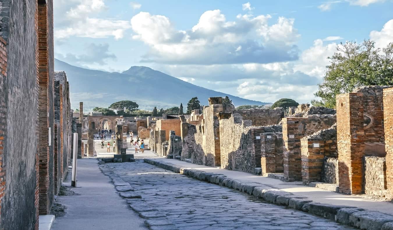 Rua de paralelepípedos em Pompéia com ruínas de edifícios e o Monte Vesúvio ao fundo.