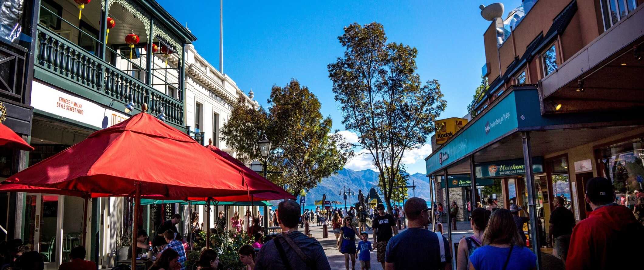 Pessoas caminham pelas ruas da histórica Queenstown, Nova Zelândia, com um lago e montanhas ao fundo