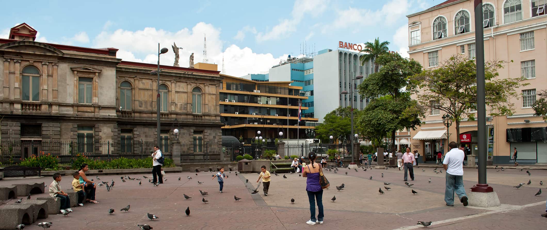 Pessoas caminham por uma praça central cheia de pombos em San José, Costa Rica