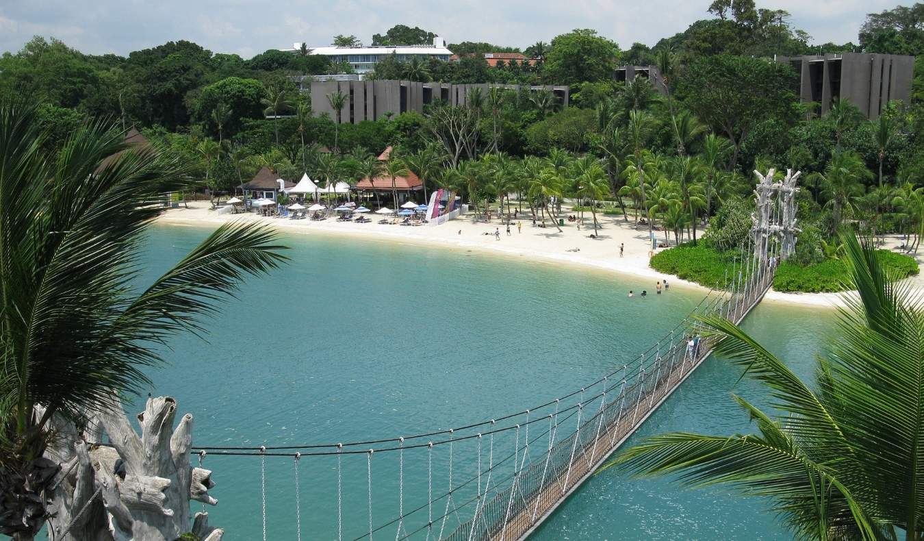 Ponte suspensa para pedestres que leva a uma praia arenosa com palmeiras e pequenas cabanas em Sentosa, Singapura