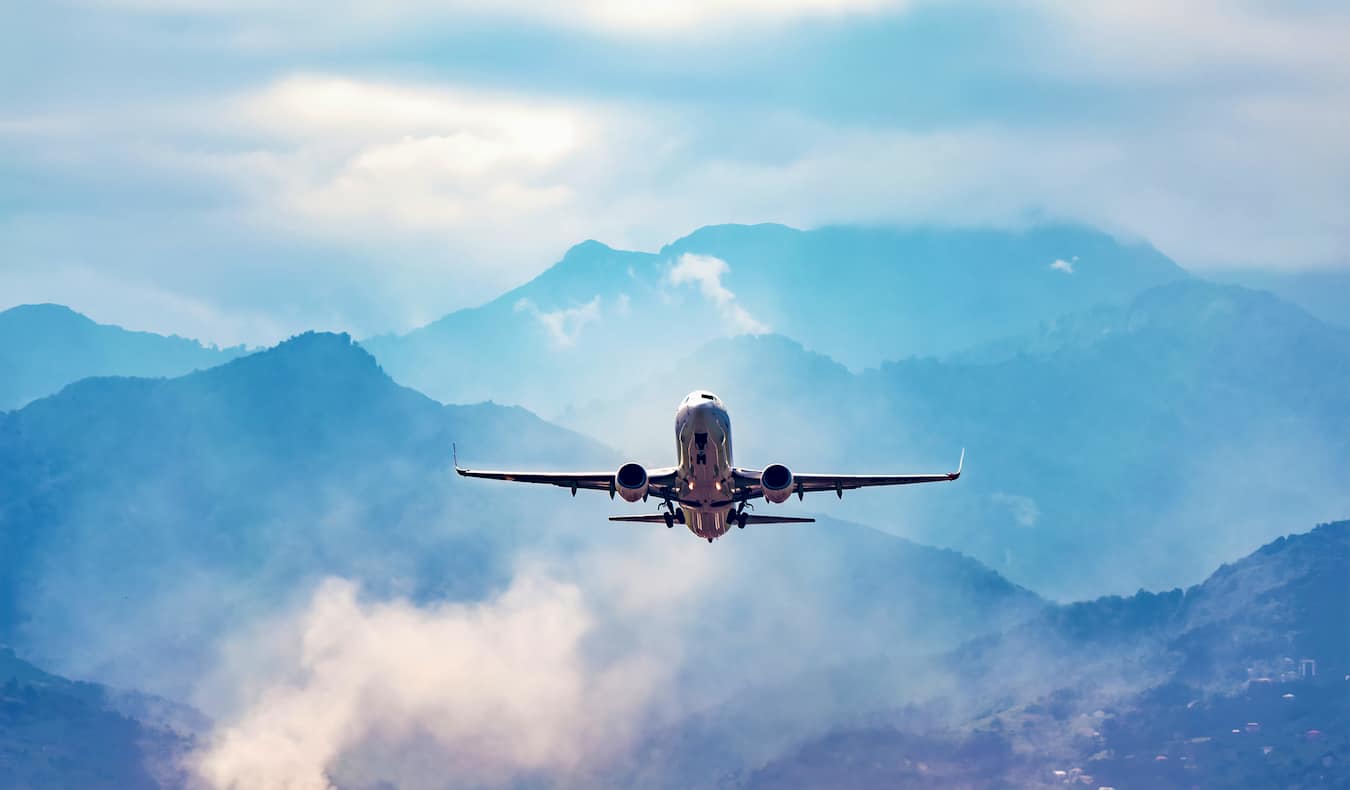Um avião comercial solitário voa através de um céu azul brilhante com montanhas ao longe.
