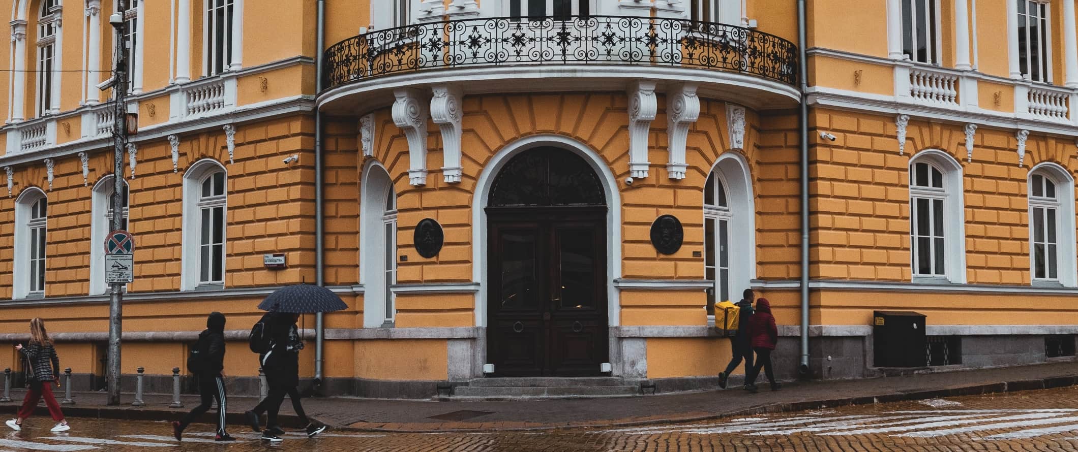 Pessoas caminham pela rua na chuva, passando por um prédio amarelo brilhante em Sófia, Bulgária