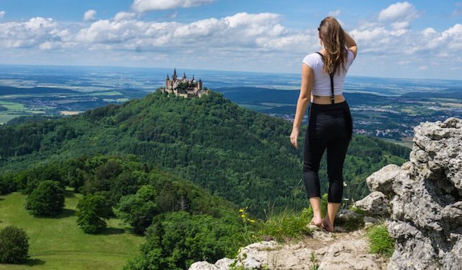 Christine olha para um belo castelo à distância no topo de uma colina verde exuberante na Europa