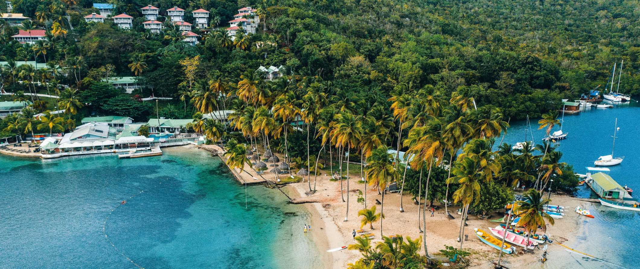 Vista do drone para uma pequena baía de turquesa com barcos ao longo da praia estabelecidos por palmeiras e casas empoleiradas nas magníficas encostas das colinas de Saint-Lucia