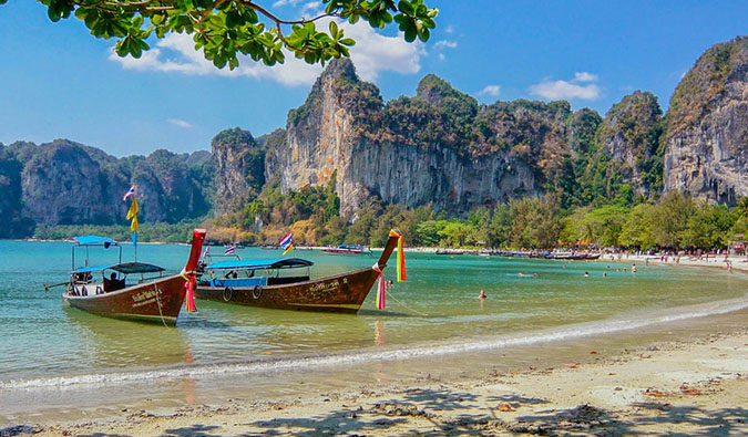 Barcos longtail vermelhos aproximam-se de uma praia pitoresca com pedras ao fundo, Tailândia