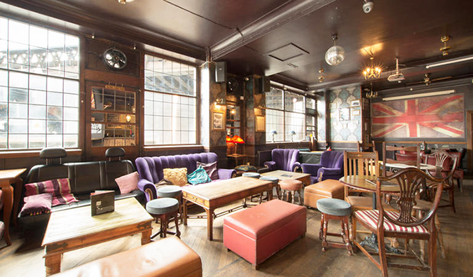 Uma sala comum acolhedora e espaçosa com muitas mesas e cadeiras de madeira, sofás roxos e uma grande bandeira britânica na parede no albergue The Walrus, Londres