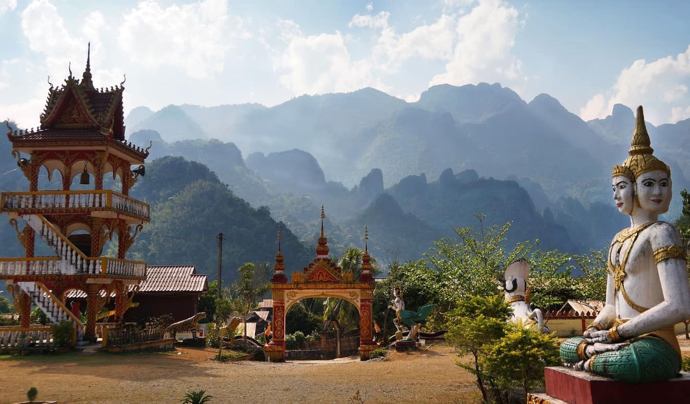 Estátua budista, pagode e portões vermelhos no cenário de montanhas em Wang Veenga, Laos
