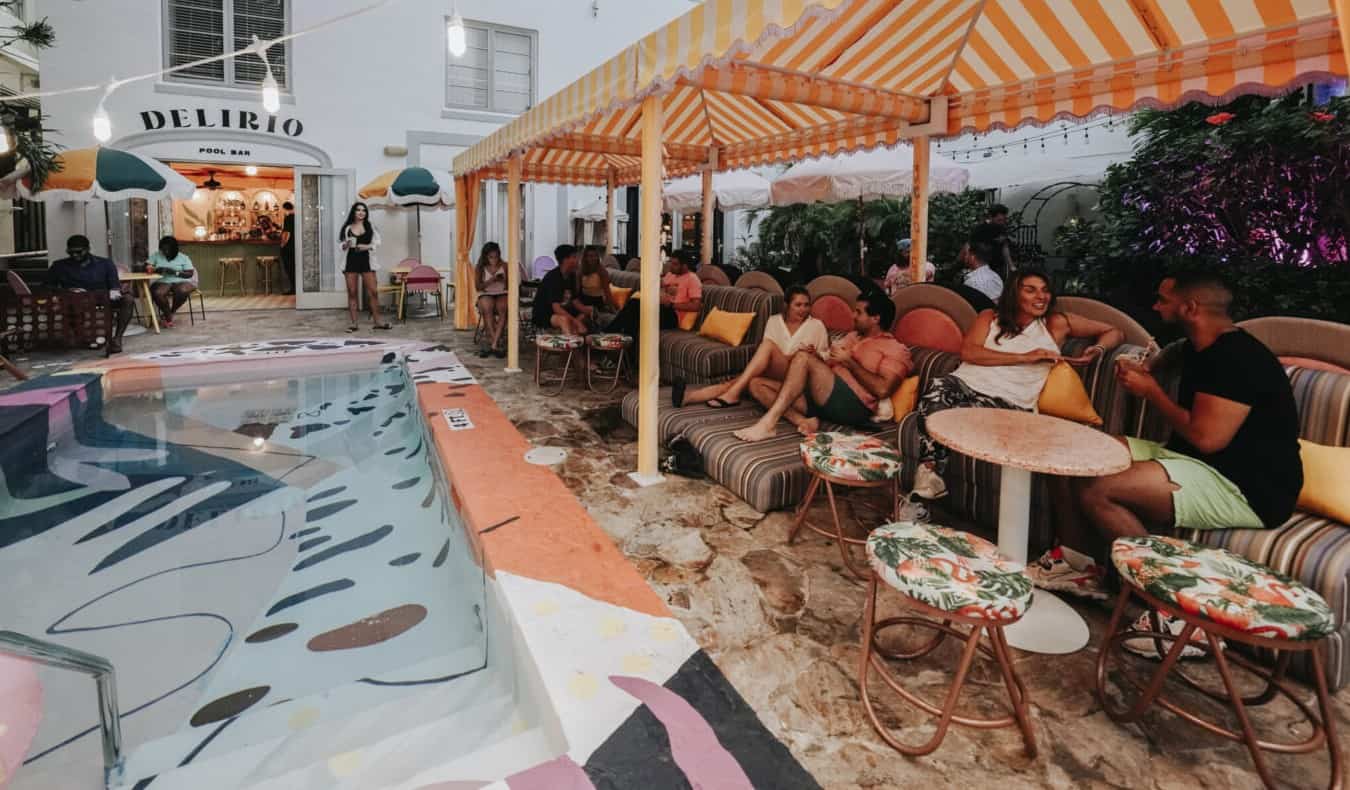As pessoas estão sentadas em pequenas mesas próximas a uma piscina aberta com azulejos mult i-coloridos no Viajero Hostel em Miami, Flórida
